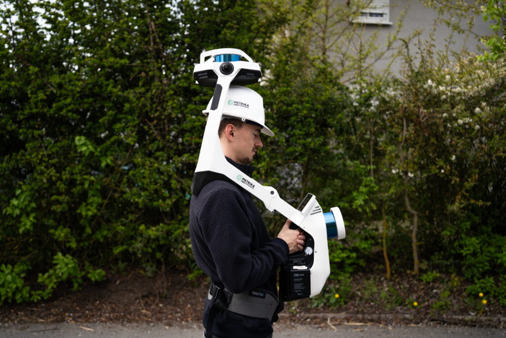 30 Mitarbeiter mit Helm trägt einen mobilen Laserscanner auf einem Weg vor einer Hecke