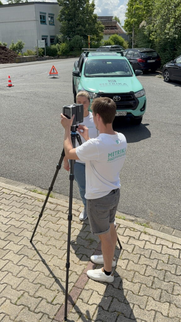 Zwei Mitarbeiter richten einen Laserscanner auf dem Stativ ein, dahinter der Firmenwagen