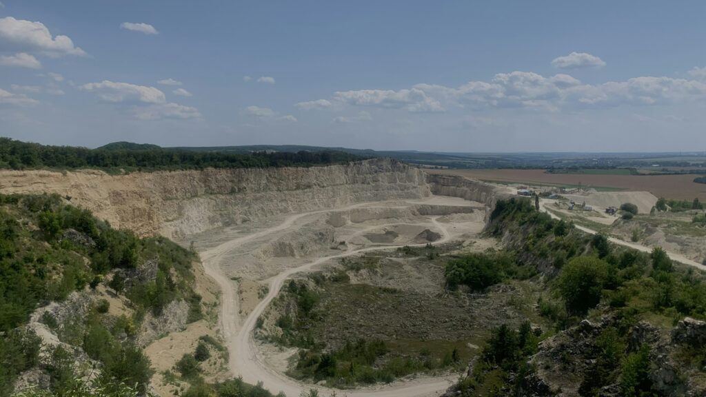 Steinbruch im Ueberblick: abgestufte Abbauwaende, Fahrwege und Halden, dahinter die offene Feldflur.