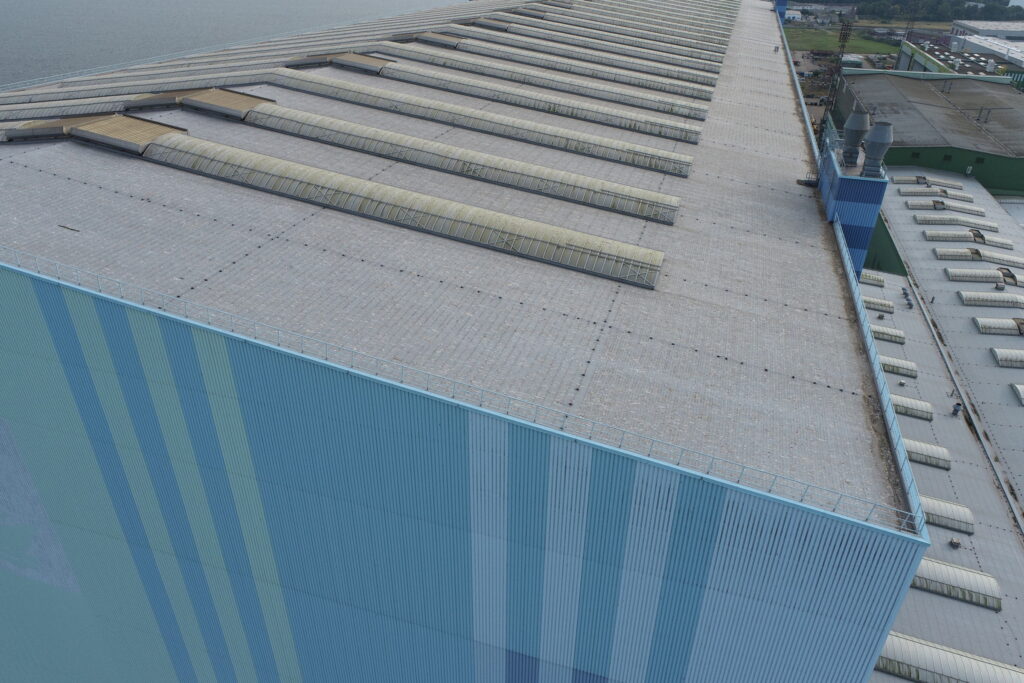 werfthalle-dachaufnahme-drohne Dach einer Werfthalle aus der Drohne: Reihen von Tonnenoberlichtern auf der Dachfläche, dahinter Hafenbecken und weitere Hallen.